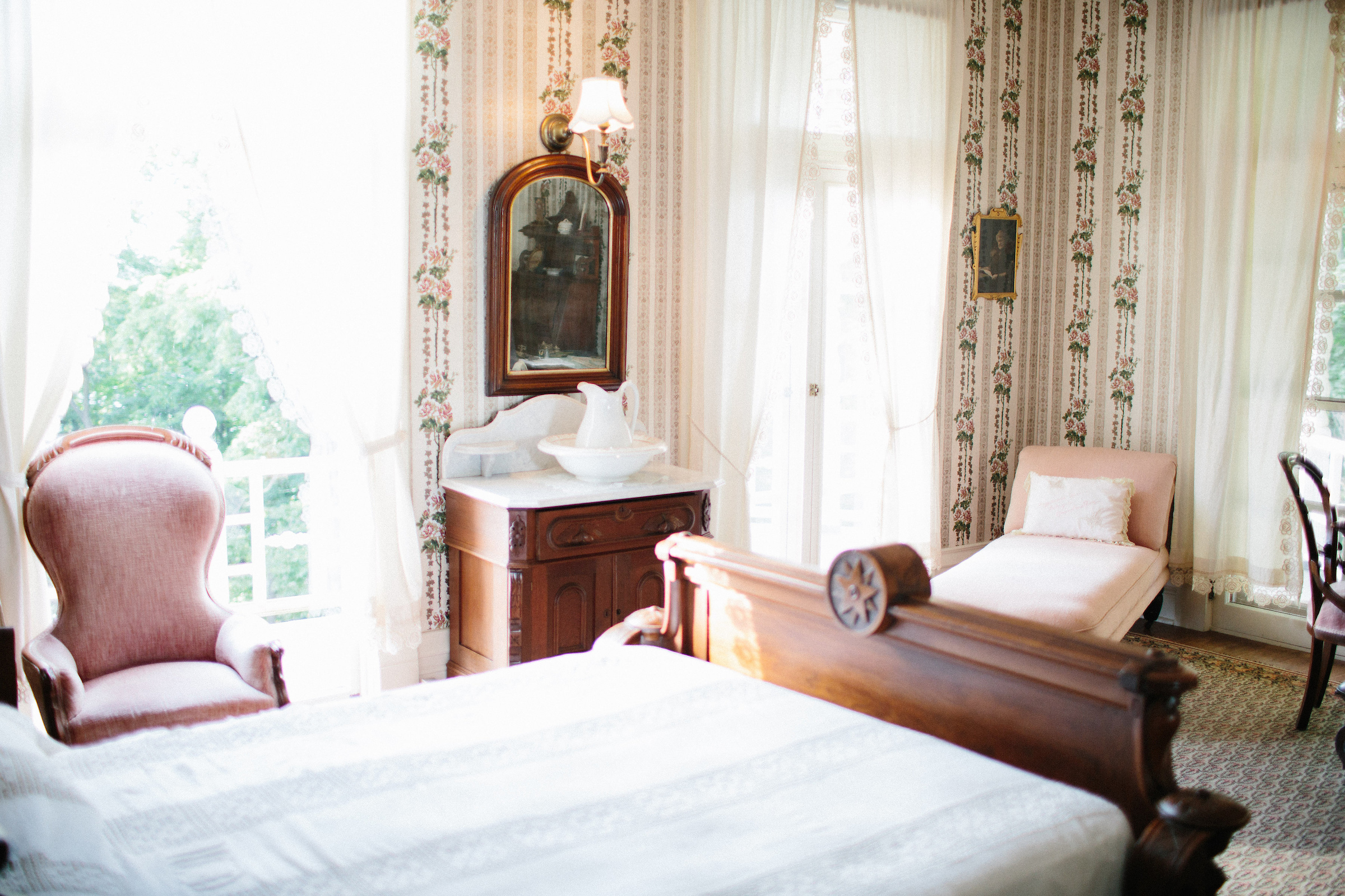 One of 13 bedrooms in the cottage, dark wood furniture, in a decorative queen anne's style. white bed spread, pink upholstered chairs, and floral wall paper with white curtains. 