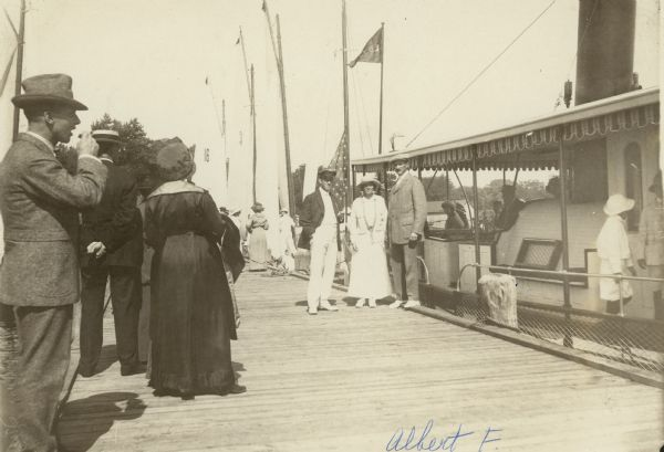 The focal point of the image is the Seipp couple and a friend. The stand on a boat dock adjacent to a docked ferry with other people waiting on the dock off to the right. 