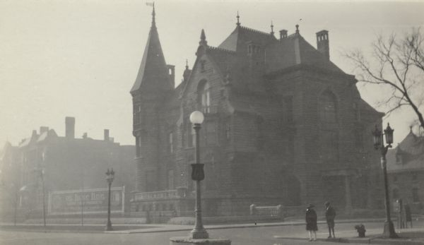 View across street towards a boy and girl standing on the street corner across from the granite mansion built for Conrad Seipp at 3300 South Michigan Avenue. The home features several chimneys and a tower with turret. There are ornamental ironwork spires on the roof. A billdboard advertising "De Luxe Line" stationary products stands incongruously in a vacant lot next door on the left. The house was designed by Adolph Cudell and was completed in 1888.